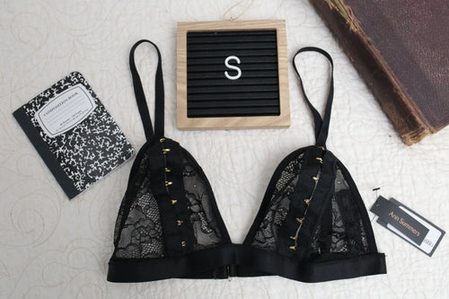 A black lace open-cup bra with adjustable straps, sheer lace cups, and a hook and eye peephole detail, displayed on a flat surface with a notebook and a letterboard.