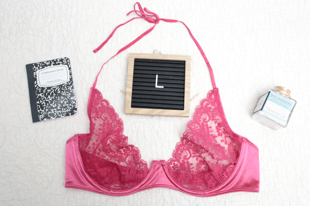 An overhead shot of a hot dark pink unlined lace bra with a halter neck design, displayed alongside a black notebook, a perfume bottle, and a letterboard with the letter 'L' on it.