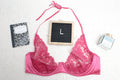 An overhead shot of a hot dark pink unlined lace bra with a halter neck design, displayed alongside a black notebook, a perfume bottle, and a letterboard with the letter 'L' on it.