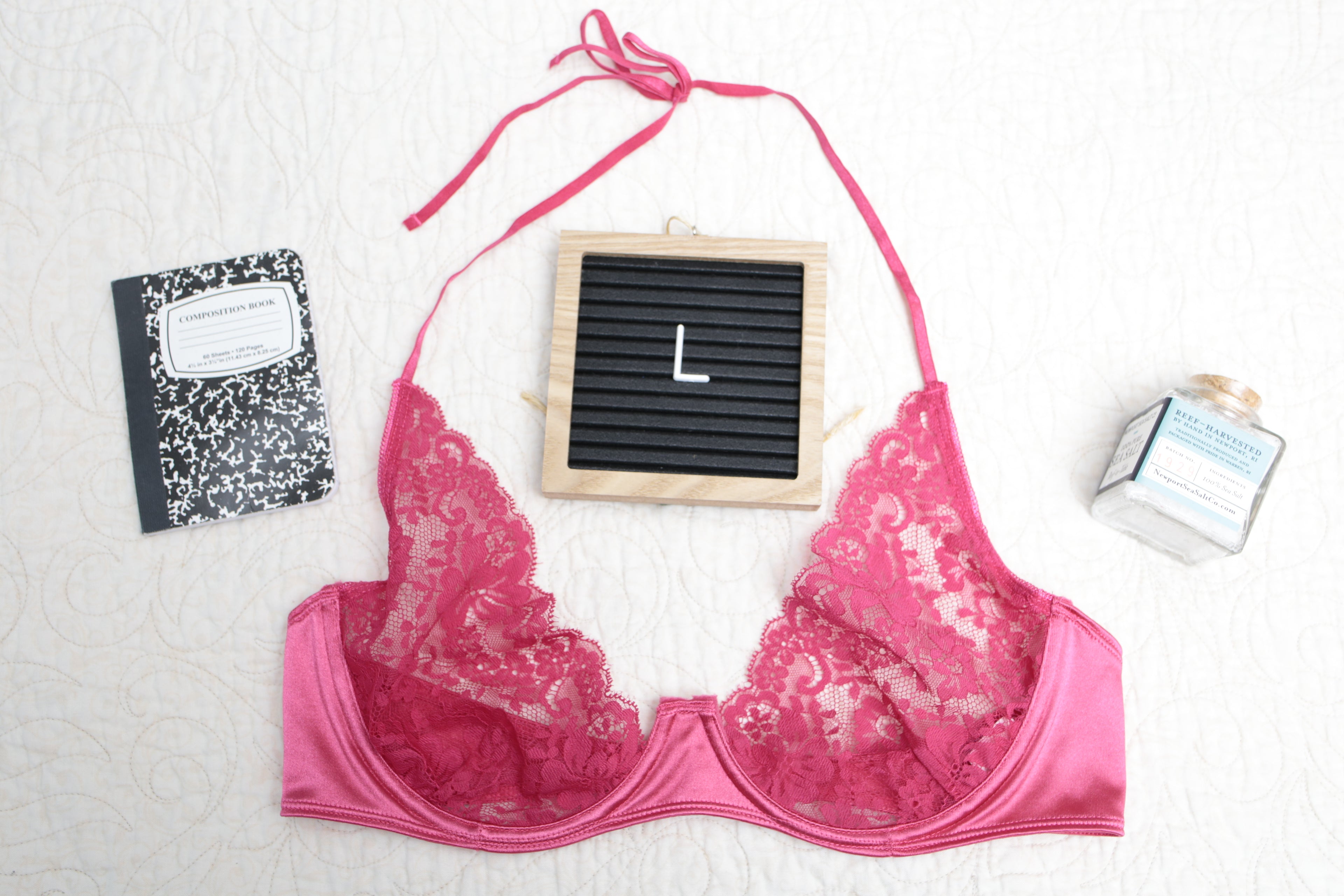 An overhead shot of a hot dark pink unlined lace bra with a halter neck design, displayed alongside a black notebook, a perfume bottle, and a letterboard with the letter 'L' on it.
